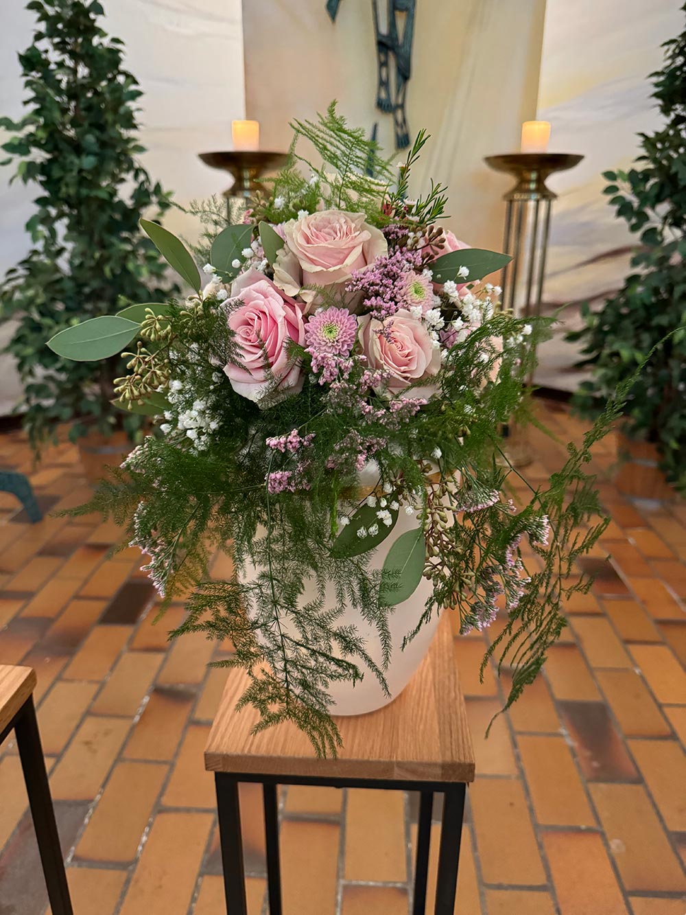 Ein Blumenstrauß mit rosa Rosen und zartem Grün steht auf einem Holzpodest