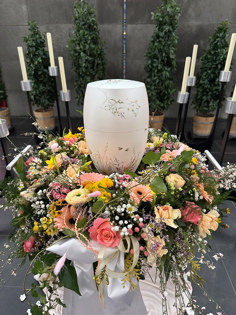 Eine weiße Urne mit floralen Mustern steht auf einem bunten Kranz aus frischen Blumen