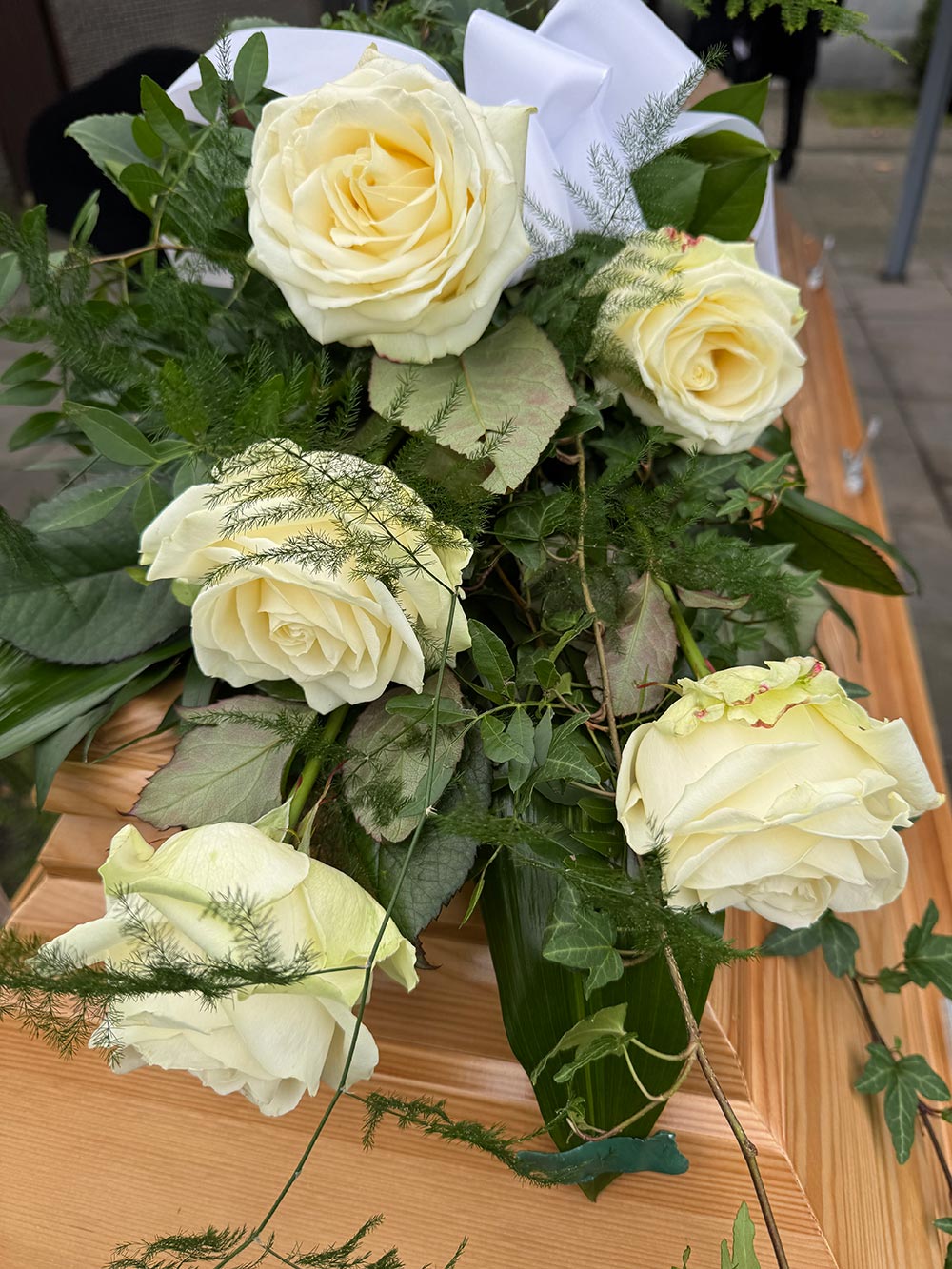 Sechs cremefarbene Rosen mit grünen Blättern liegen auf einem hellen Holzsarg