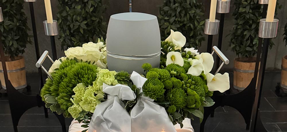 Graue Urne mit weißen und grünen Blumen sowie Kerzen im Hintergrund aufgebahrt