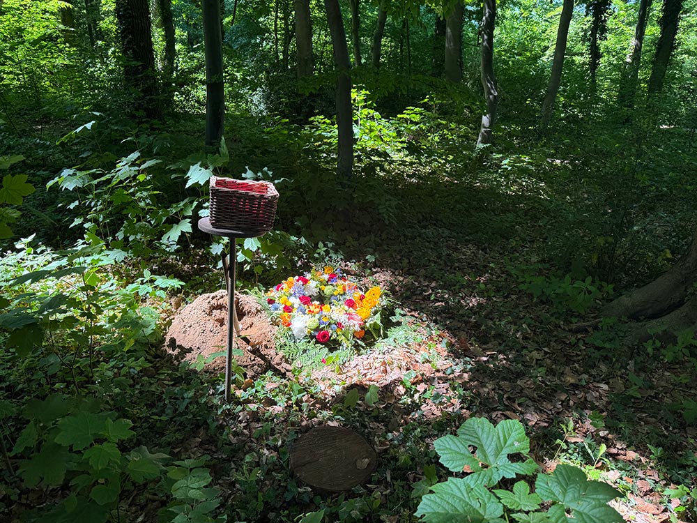 Ein frisches Grab im sonnigen Wald mit bunten Blumen und einem roten Korb auf Ständer