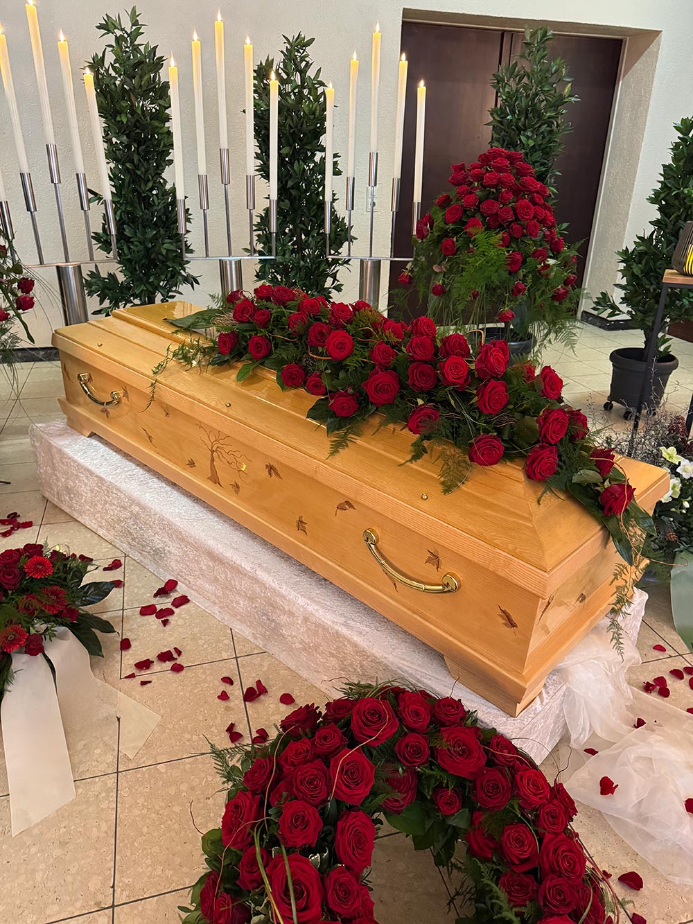 Ein geschlossener heller Sarg mit vielen roten Rosen liegt in einem feierlich geschmückten Raum