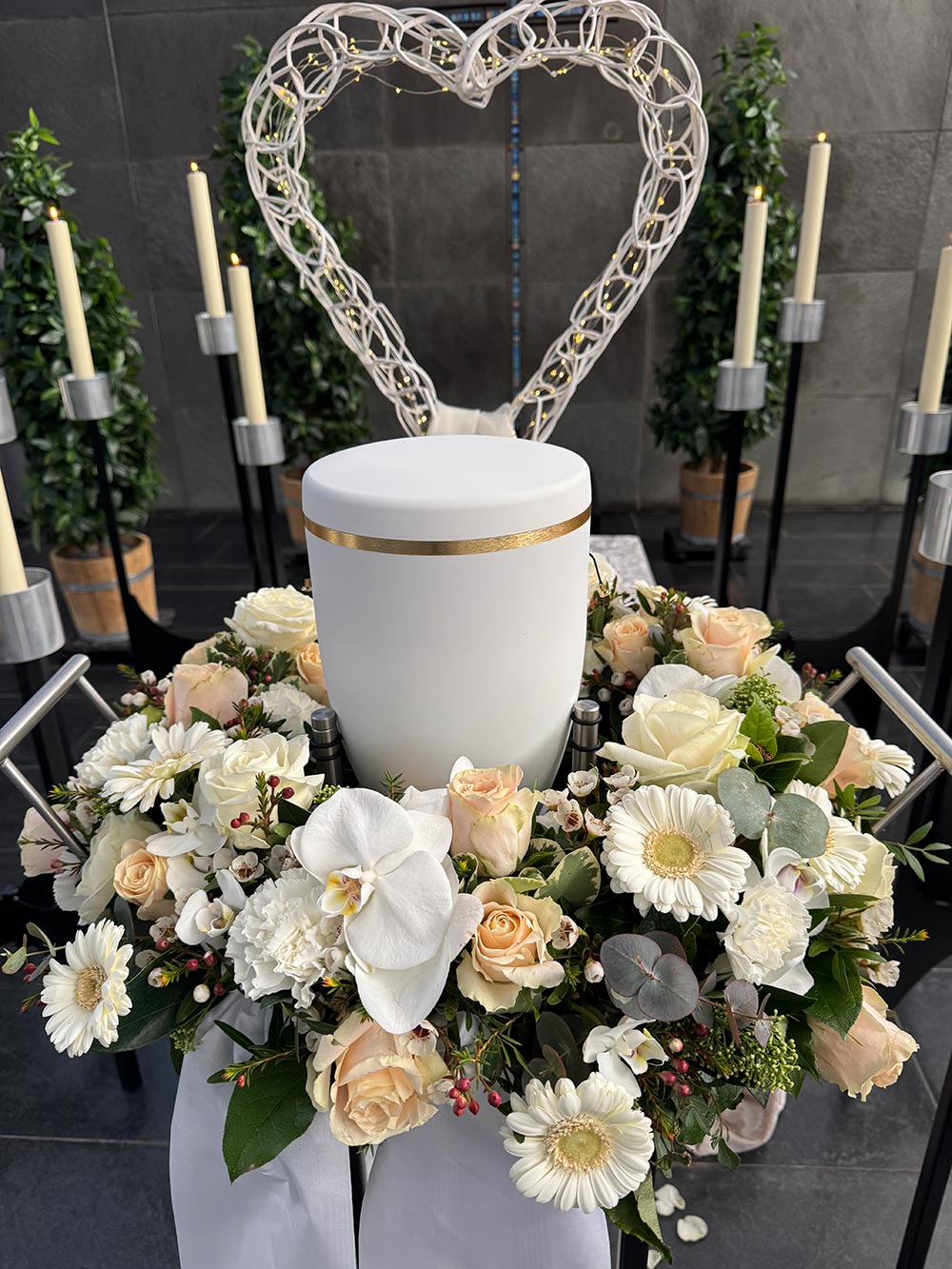 Eine weiße Urne steht inmitten heller Blumen und vor einem Herz aus Draht