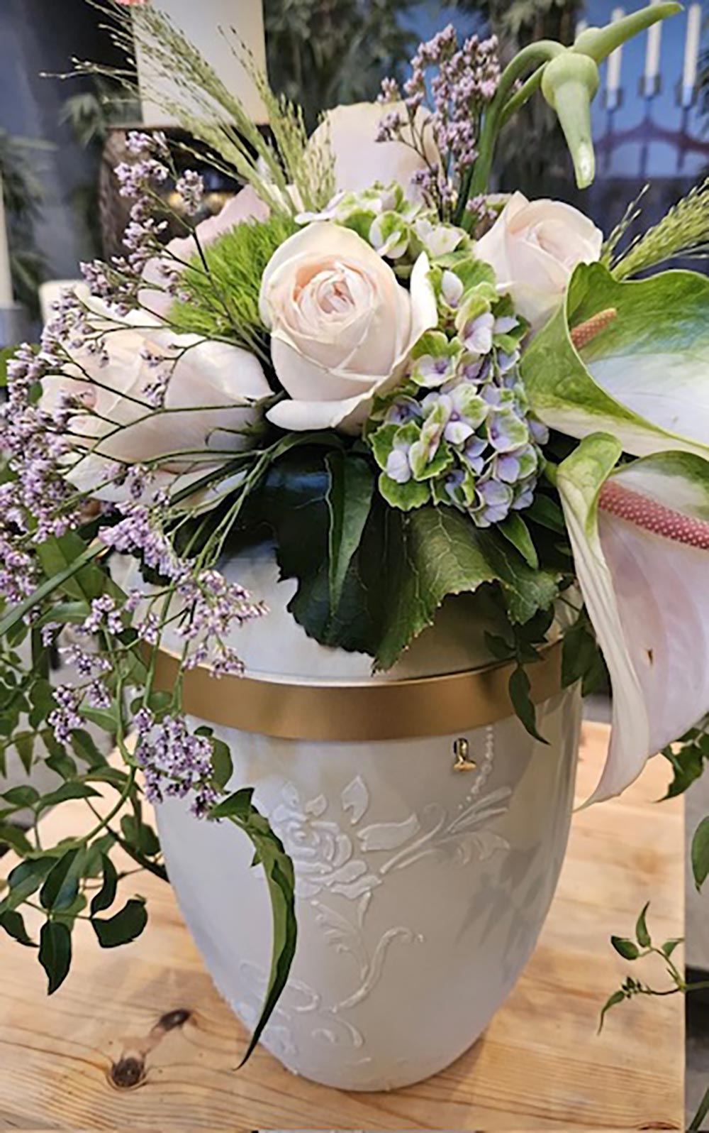 Eine helle Urne mit goldenem Band wird von einem zarten Blumenarrangement geschmückt