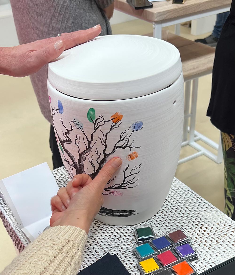 Zwei Frauen zeigen mit neugierigen Blicken auf eine bunte Urne mit Baum-Motiv